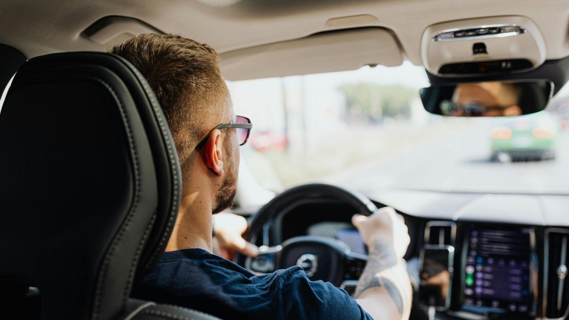 A person with glasses driving a car, seen from behind. The dashboard displays navigation and a blurred road is visible through the windshield.