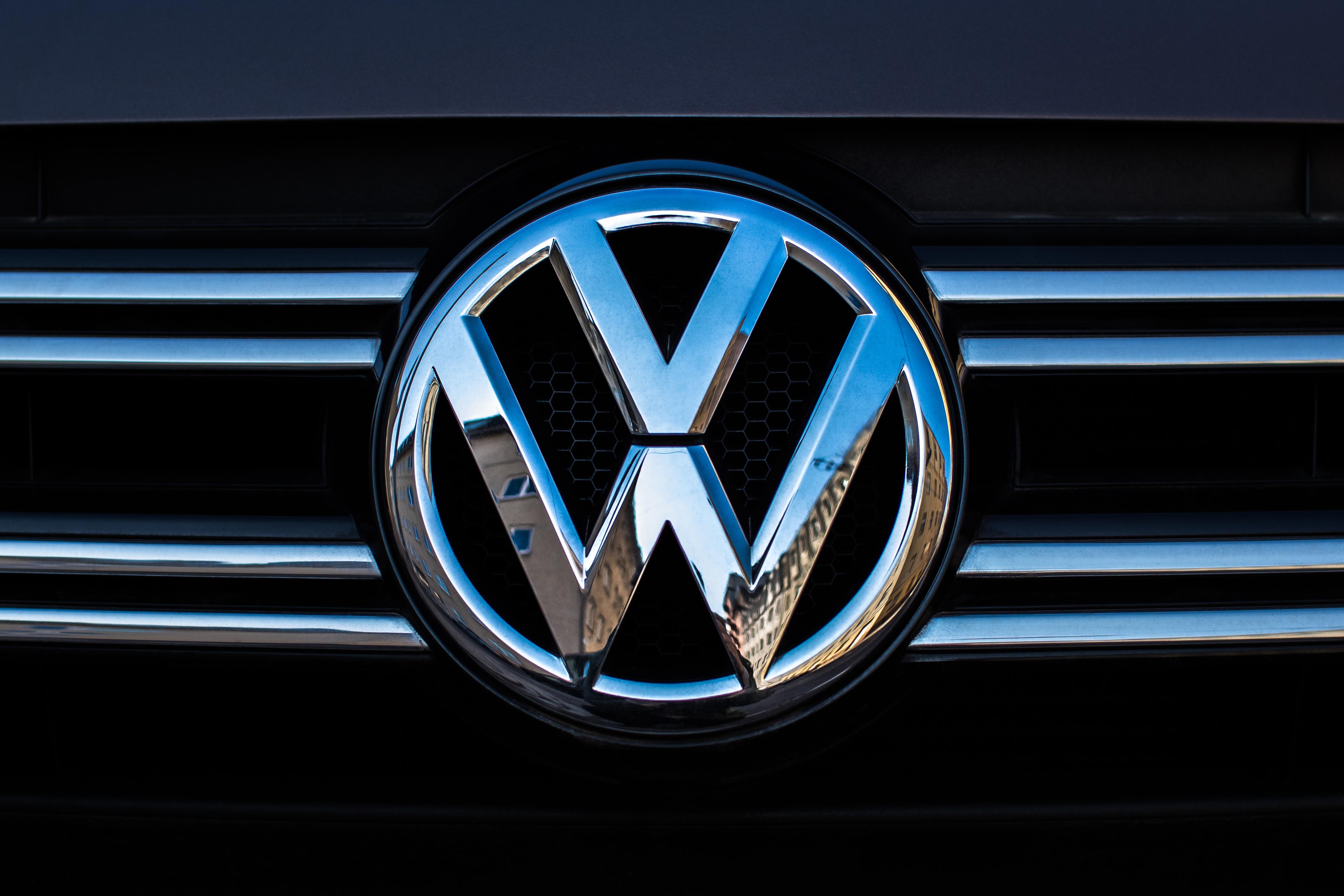 Close-up of a Volkswagen logo on a car grille with chrome horizontal bars, reflecting nearby buildings.