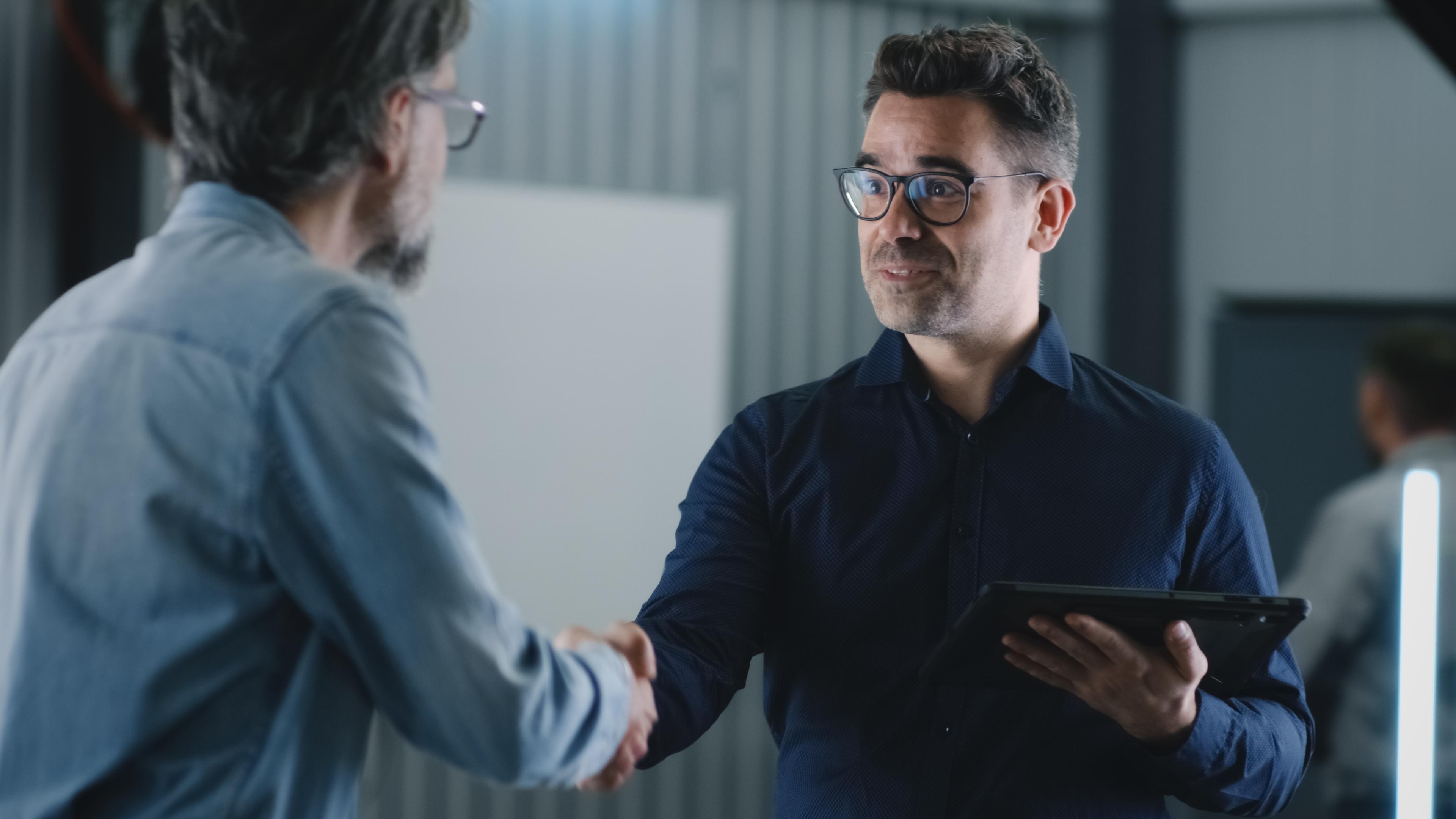 Two men shaking hands indoors; one holds a tablet and wears glasses, both in casual shirts, with a blurred background.