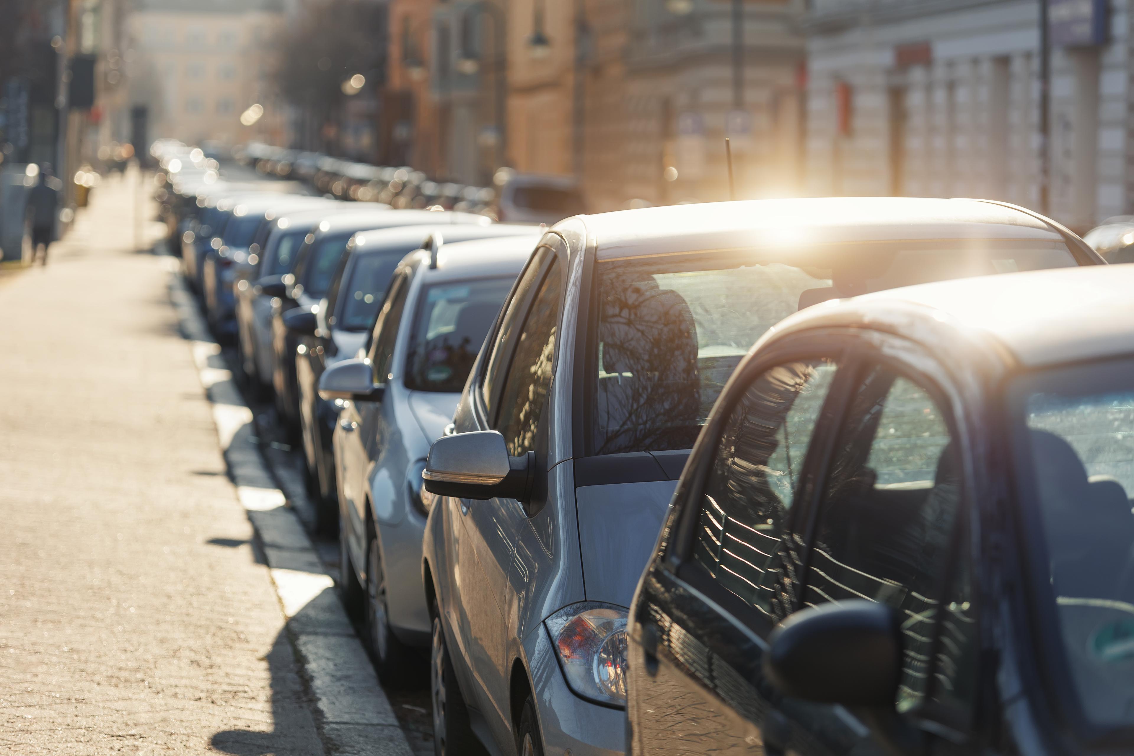 A row of parked cars lining a sunlit urban street, with sunlight reflecting off the car surfaces.