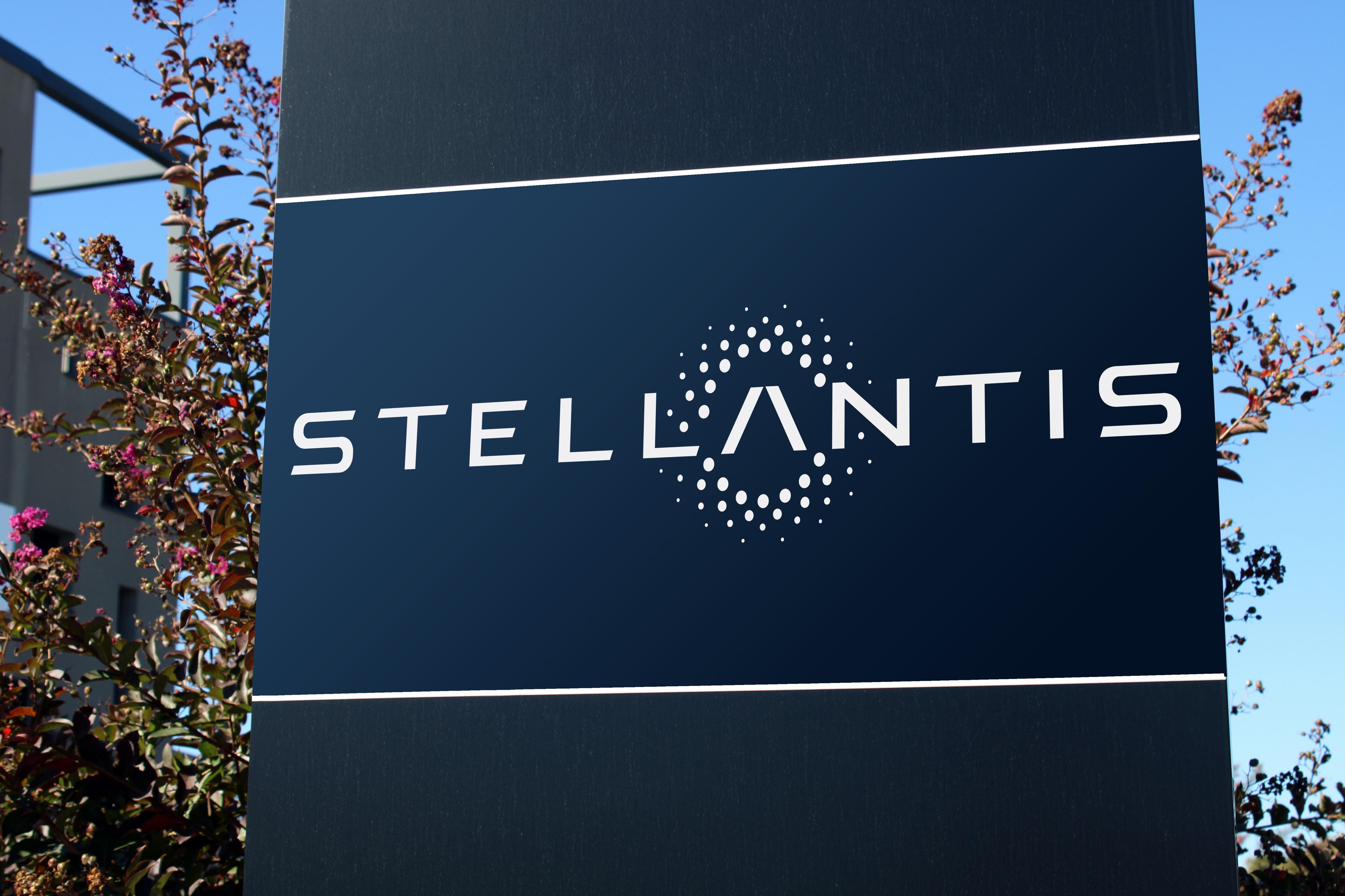 Sign with Stellantis logo on a dark blue background, featuring circular dot patterns, with plants and a clear sky in the background.