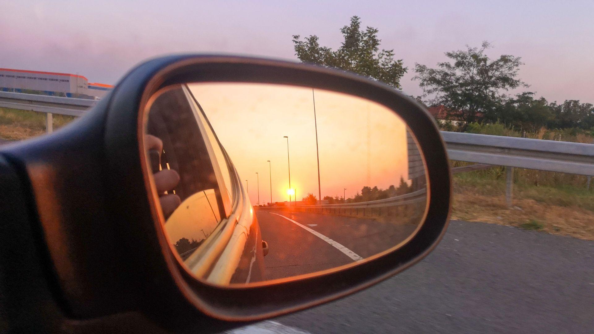 Rearview mirror reflecting a sunset over a highway with streetlights, trees, and guardrails.