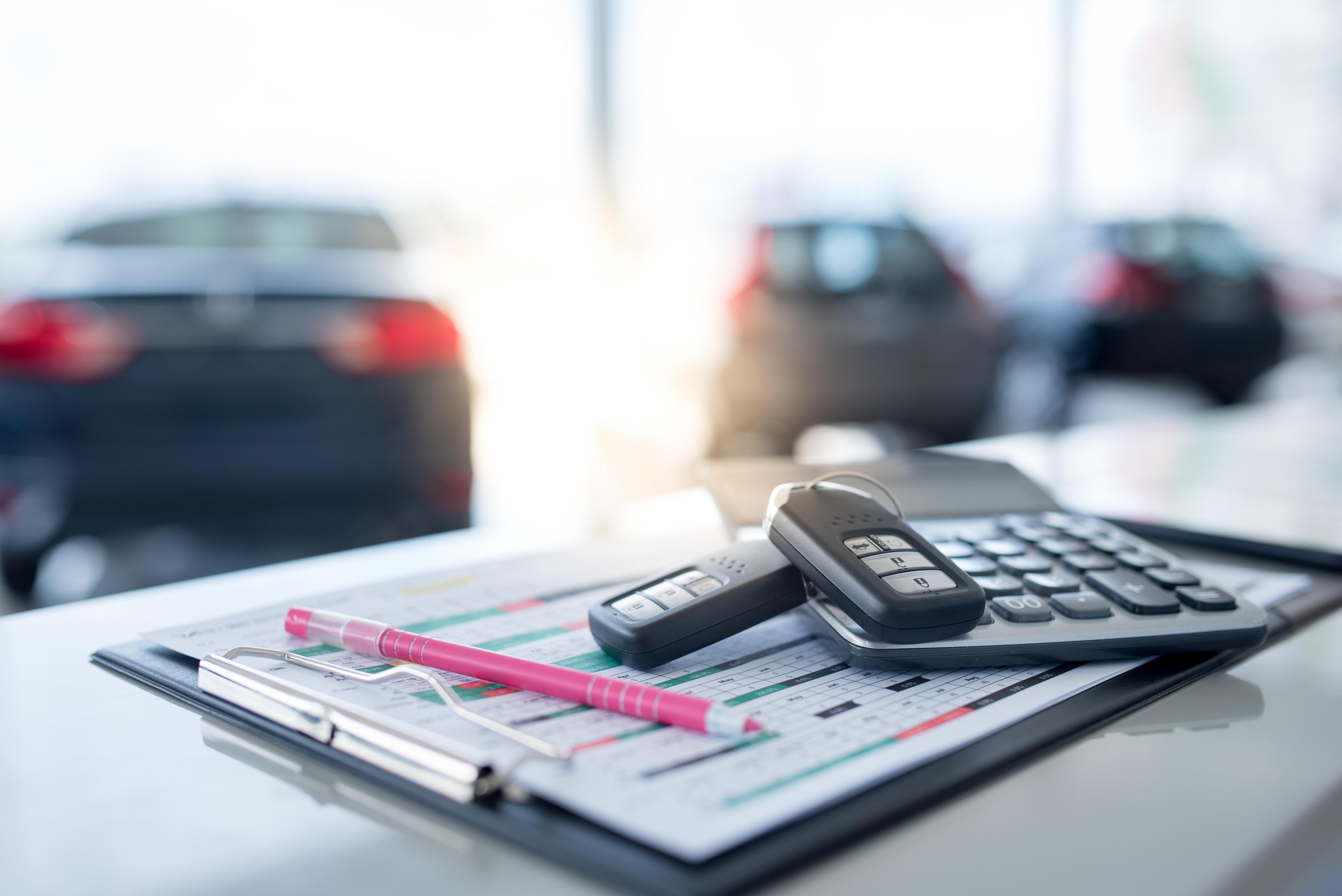 lost-of-advised-repairs-next-to-car-keys-next-to-calculator Car keys, a calculator, and a pink pen on a clipboard with papers in a car showroom, blurred vehicles in the background.