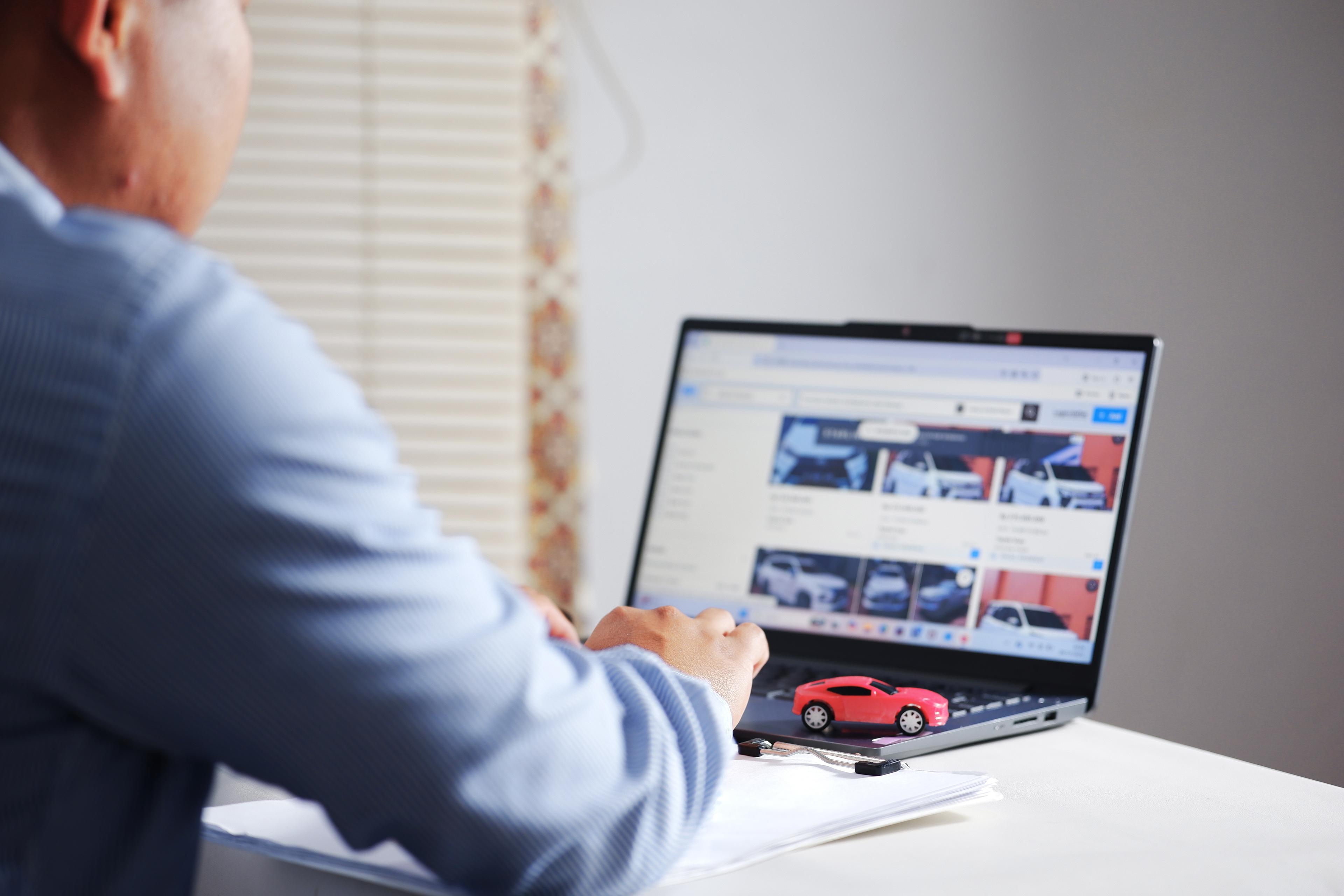 male-customer-using-ai-to-compare-vehicle-options Person browsing car listings on a laptop, with a small red car model beside it.