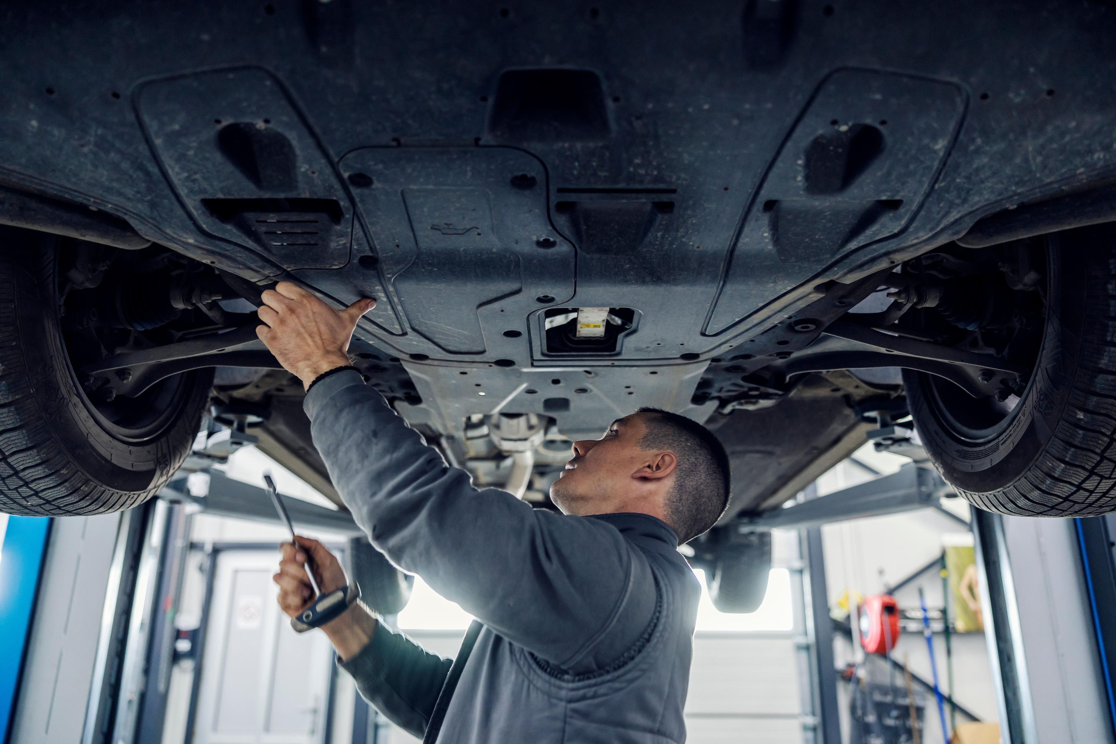 Mechanic inspecting the underside of a car on a lift, holding a tool in a well-lit garage.