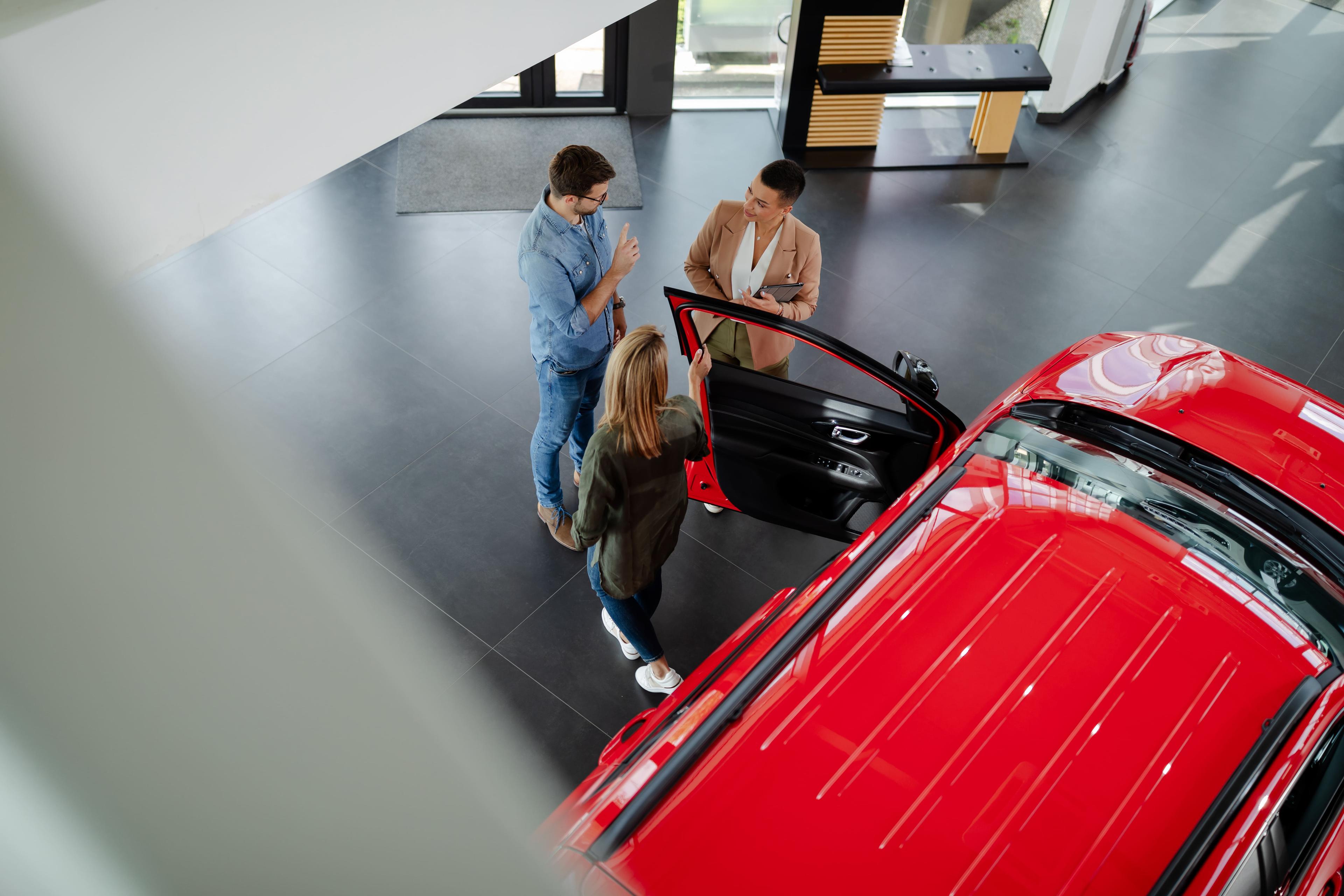 Overhead view of two customers with a salesperson discussing a red car inside a showroom. One customer holds the open car door.